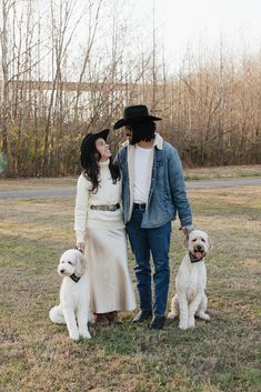 a man and woman standing next to two dogs in a field with trees behind them