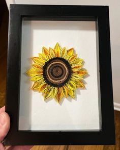 a hand holding up a framed artwork with a yellow sunflower on the inside of it