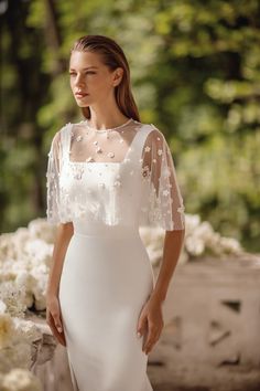 a woman standing in front of flowers wearing a white wedding dress with sheer sleeves and beading