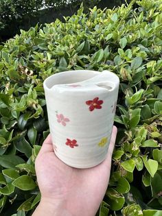 a hand holding a white cup with red flowers on it in front of some bushes