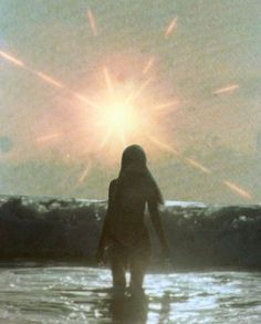 a person standing in the water with their back to the camera, looking at the sun