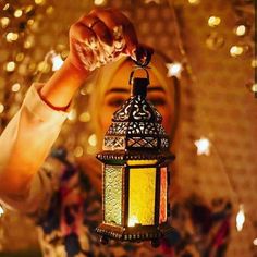 a woman holding a lit lantern in front of her face with lights on the wall behind her