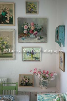 several paintings on the wall above a table with flowers in vases and other items