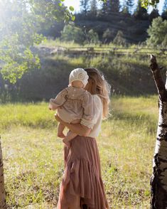 a woman holding a baby in her arms while standing next to a tree and grass field