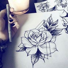 a person holding a pen and drawing on paper with flowers in the background, next to a candle