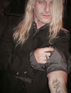 a man with long blonde hair wearing a black shirt and holding his arm around his chest