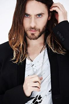 a man with long hair and blue eyes wearing a black blazer, white t - shirt and silver necklace