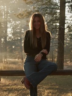 a woman sitting on top of a wooden bench in front of a forest filled with trees