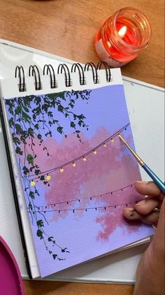 a person is holding a pen and drawing on a notebook with string lights in the background