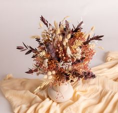 an arrangement of dried flowers in a white vase on a yellow cloth covered bed spread