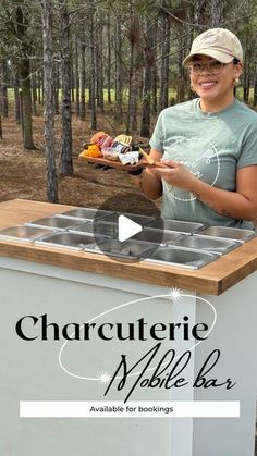 a woman standing in front of a mobile bar with food on the counter and trees behind her