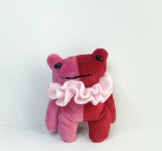 a pink and red teddy bear with a ruffle around it's neck sitting against a white wall