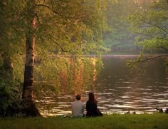 two people are sitting on the grass next to a body of water with trees in the background