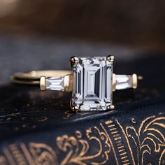 an emerald - cut diamond ring sits on top of a book with gold trimmings