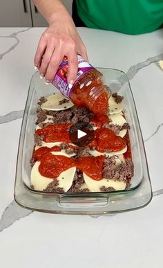 a person pouring sauce on some food in a glass baking dish with meat and cheese