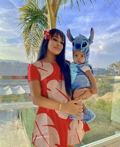 a woman holding a baby in front of a palm tree and looking at the camera