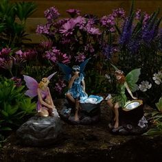 three fairy figurines sitting on top of a rock in front of purple flowers