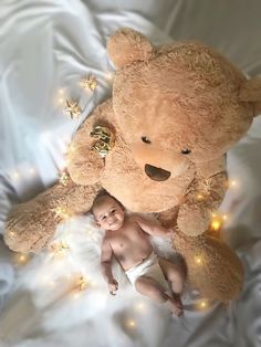 a baby laying next to a large teddy bear on a bed with lights all around it