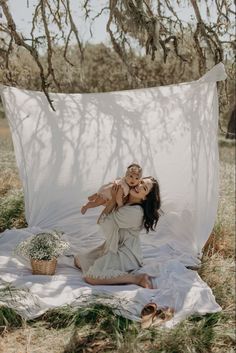 a woman holding a baby in her arms while sitting on a blanket under a tree