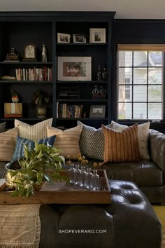 living room with dark blue painted bookshelf built into the wall. a grey sofa arranged with many neutral throw pillows to lighten the room. Pillows For A Black Leather Couch, Pillows For Dark Brown Leather Couch Ideas, Styling A Dark Leather Couch, Dark Dramatic Living Room, Grey Leather Couch Pillow Ideas, Dark Brown Leather Couch Pillow Ideas, Dark Blue And Beige Living Room, Dark Blue Furniture Living Room, Throw Pillows On Leather Couch