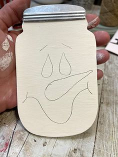 a hand holding a jar with a face drawn on it's side and the lid painted white