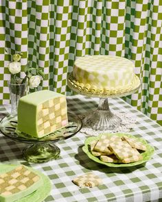 a table topped with cakes and desserts covered in frosting next to green checkered curtains