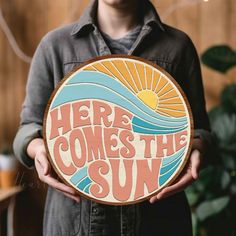 a man holding up a wooden sign that says here comes the sun