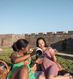 two women are sitting on the grass and having drinks