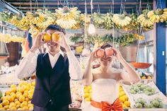 two people standing in front of fruit stand with bananas and oranges hanging from the ceiling