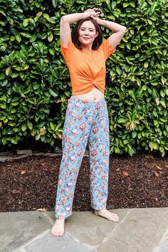 a woman standing in front of a hedge wearing pajamas and an orange shirt with her hands on her head