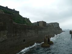 an old stone wall next to the ocean with buildings on top and cliffs in the background