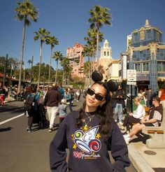 a woman wearing mickey ears standing in the middle of a street with buildings and palm trees