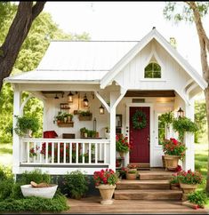 a small white house with potted plants on the front porch and red door surrounded by greenery