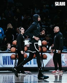 two men in black hoodies are walking on the court with basketballs behind them