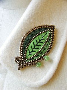 a green leaf brooch sitting on top of a white shirt with beaded trim