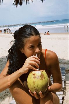 a woman sitting on a beach with a coconut in her hand and a straw in her mouth