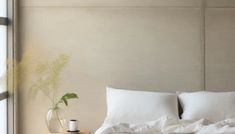 a bed with white sheets and pillows next to a plant on a nightstand in front of a window