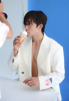 a man in a white suit eating something with his shirt open while another person looks on