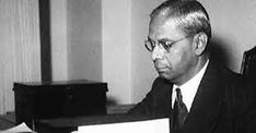 black and white photograph of man in suit sitting at desk with papers on table next to him