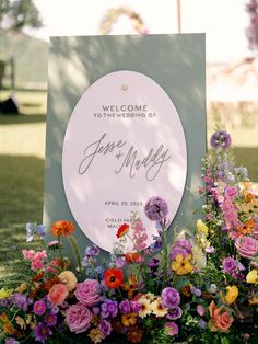 a welcome sign surrounded by colorful flowers and greenery in the grass at a wedding