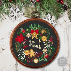 a cross stitch christmas ornament hanging from a wooden frame with pine cones, holly and bells