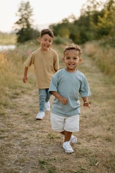 two young boys running down a dirt path together in the grass, one boy is smiling at the camera and the other has his hand on his hip