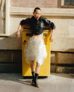 a woman standing in front of a yellow machine wearing a white skirt and black jacket