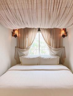 a white bed sitting under a window next to a curtained window with two lamps on each side