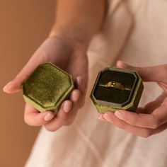 two hands holding an open ring box with a gold wedding band in it and another person's hand wearing a white dress