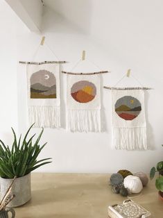 three handmade wall hangings on the wall above a table with plants and rocks