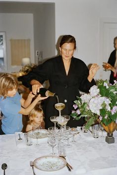 a woman pours champagne for two children at a table with flowers and wine glasses