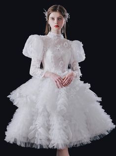 A symphony of delicate lace and cascading ruffles defines this mesmerizing wedding attire. The gown boasts a high-necked design, reminiscent of classical elegance, seamlessly blending with the voluminous puffed sleeves that add a touch of vintage charm. The bodice is a masterpiece, intricately adorned with floral embroidery, hinting at the beauty of nature. As the eyes travel downwards, they are met with layers upon layers of soft ruffles, each layer playing its part in creating a harmonious flow. The dress, with its mid-length cut, offers a fresh twist on traditional designs, ensuring the bride feels like the belle of the ball. Champagne Ball Gown, Vintage Weddingdress, Tiered Wedding Dress, Gaun Abad Pertengahan, Delicate Gown, Knee Length Wedding Dress, White Ball Gowns, Blue Ball Gowns, Ruffle Flower