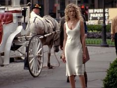 a woman in a white dress walking down the street with a horse and carriage behind her