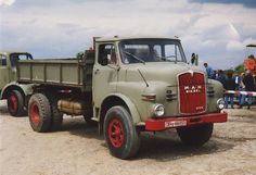 an old green truck parked on top of a dirt field next to other trucks and people